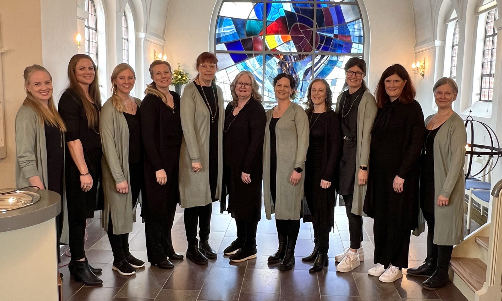 11 kvinnor i grått och svart står uppställda på rad bredvid varandra i en kyrka