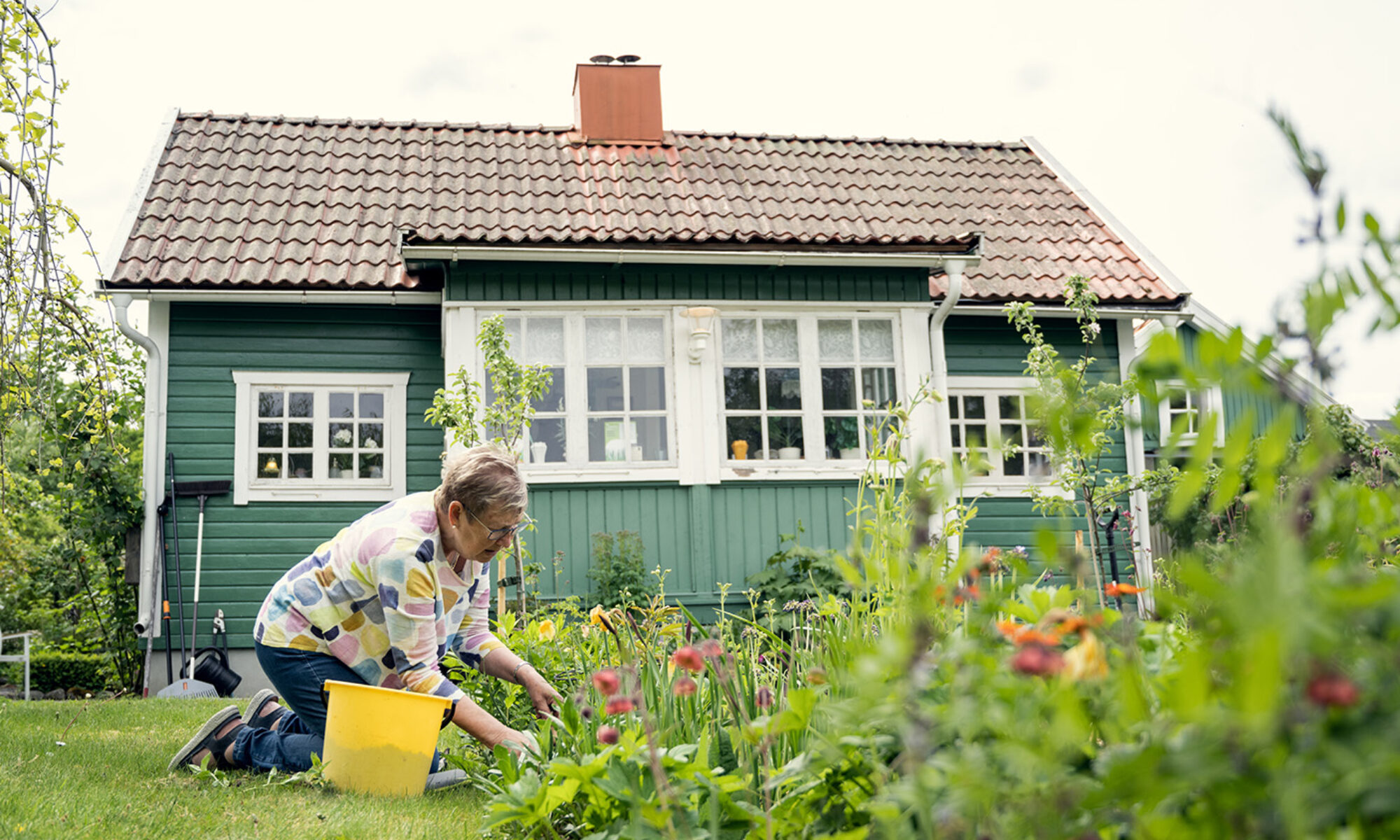 Kvinna med hink bredvid sig ligger på knä i en rabatt framför ett litet hus med spröjsade fönster